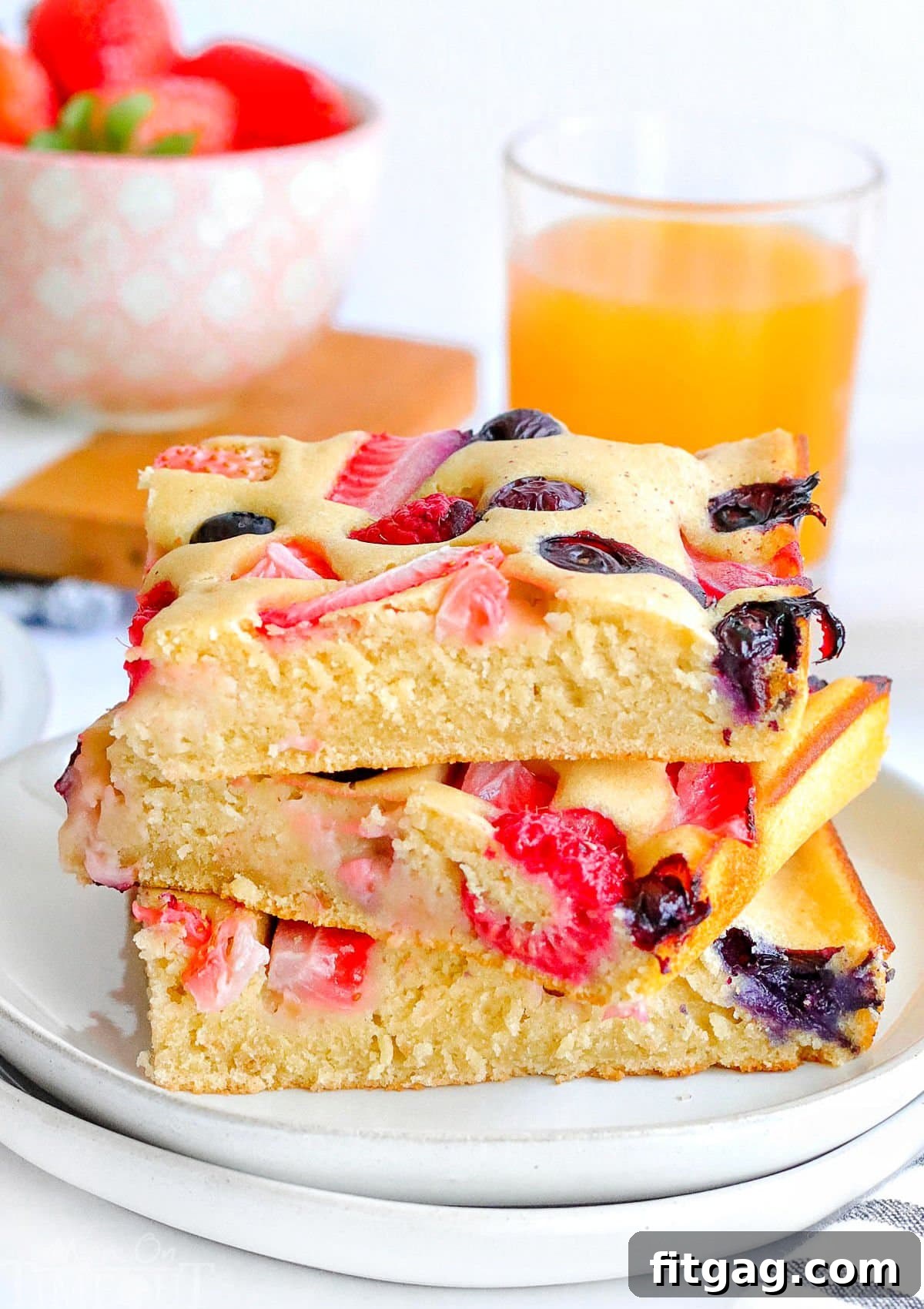 Three perfectly square berry pancakes, cut from a sheet pan, are neatly stacked on two separate white plates, creating an inviting breakfast display. A vibrant glass of orange juice sits in the background, adding a touch of freshness to the scene.