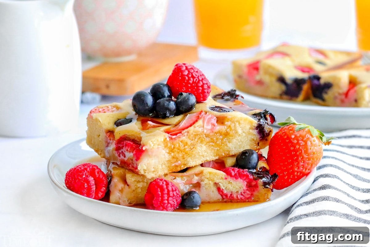 Oven Pancakes 6 Two golden-brown sheet pan pancakes, precisely cut into squares, are stacked invitingly on a clean white plate. They are artfully garnished with a vibrant assortment of fresh berries and a generous pour of glistening maple syrup.