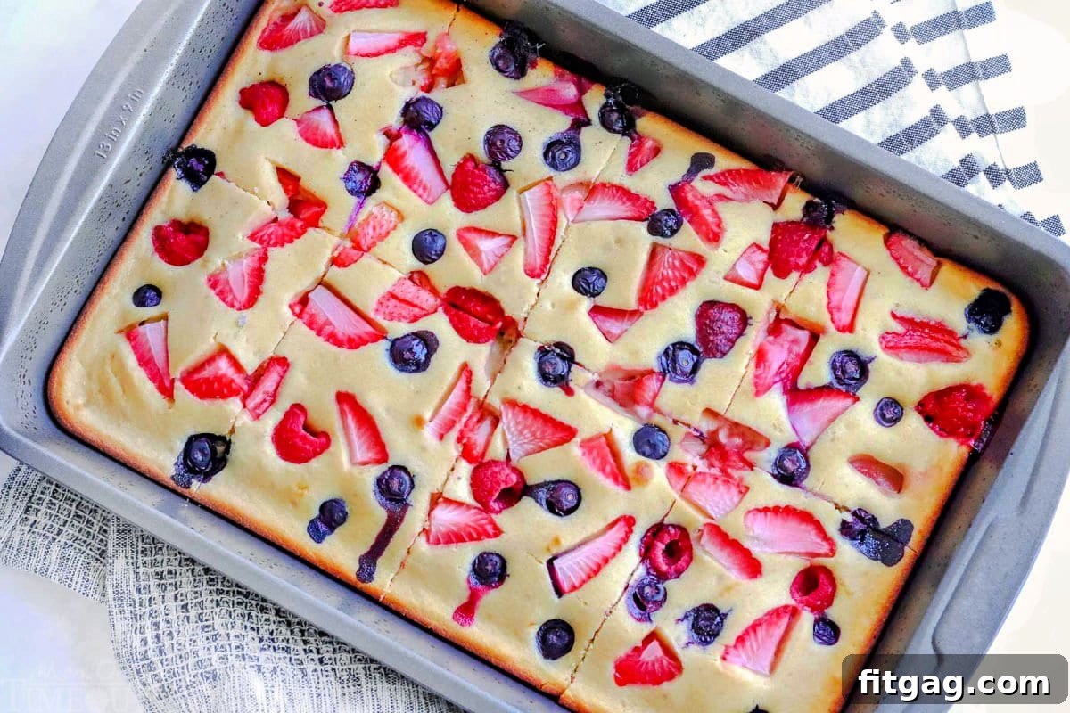 Freshly baked oven pancakes, created in a sheet pan, are lavishly topped with an array of vibrant strawberries, raspberries, and blueberries. The large pancake has been expertly cut into perfectly portioned squares, ready to be served.