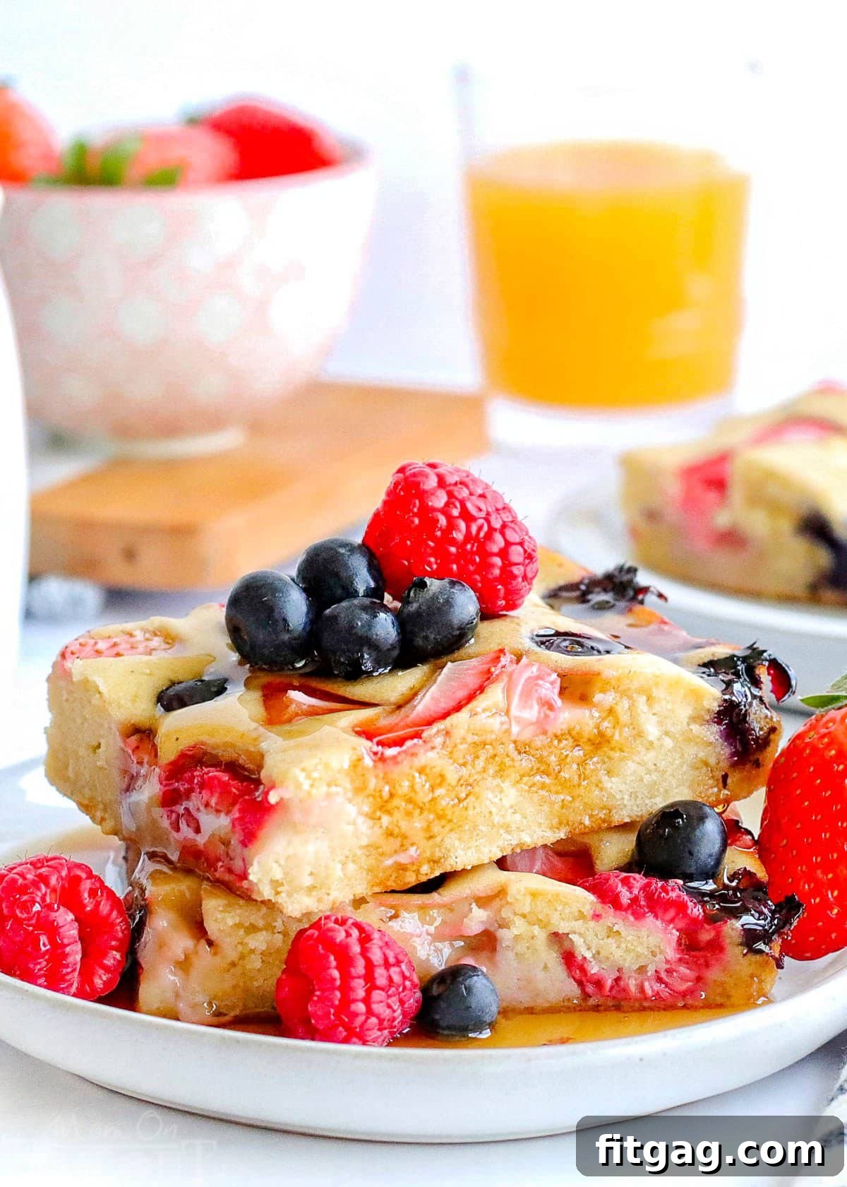 Oven Pancakes 8 Two delightful sheet pan pancakes, expertly cut into squares, are stacked appealingly on a white plate. They are beautifully adorned with a scattering of fresh, colorful berries and a luxurious cascade of golden maple syrup, ready to be enjoyed.