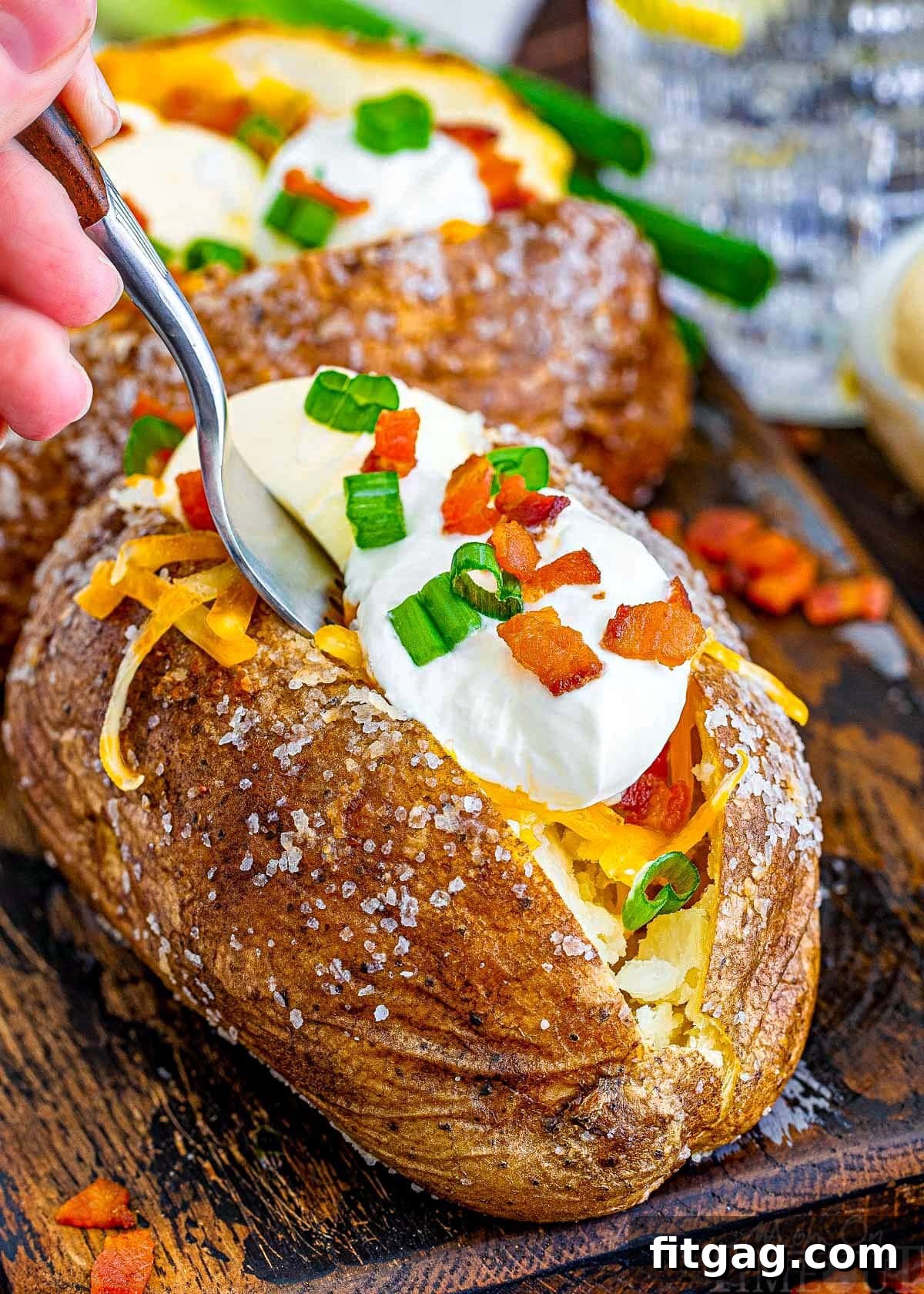 A fork is inserted into a loaded baked potato, poised to pull out a bite. The potatoes are presented on a rustic dark wood board, highlighting their inviting texture and rich toppings.