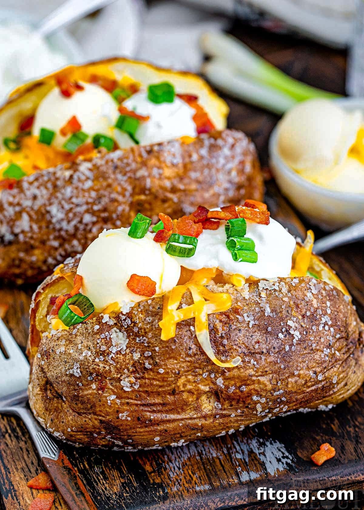 An angled shot looking down at two beautifully baked potatoes, generously topped with shredded cheese, a dollop of sour cream, and scattered bacon bits, ready to be enjoyed. The visible salt crystals on the potato skin highlight its crispy texture.