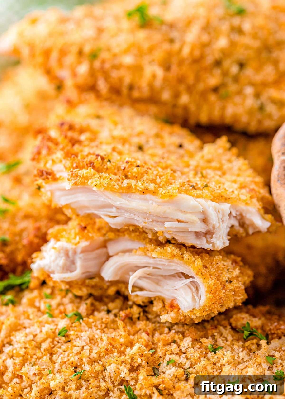 A close-up of a chicken tender torn in half, revealing its tender cooked interior and crispy breading, resting on a pile of other tenders.