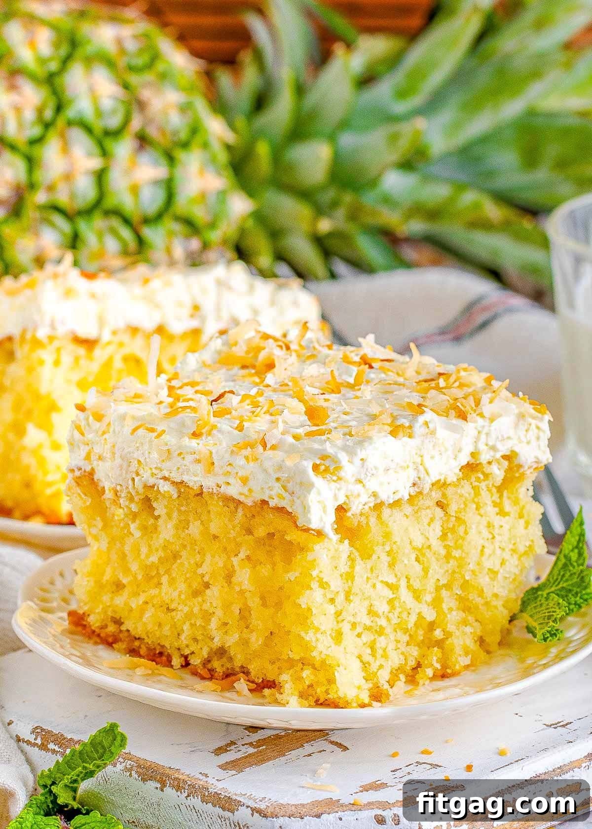A perfectly square slice of Pina Colada Cake on a white plate, elegantly frosted with white, fluffy topping and garnished with golden toasted coconut flakes. In the soft background, another slice of cake and a fresh pineapple hint at the tropical inspiration behind this exquisite dessert.