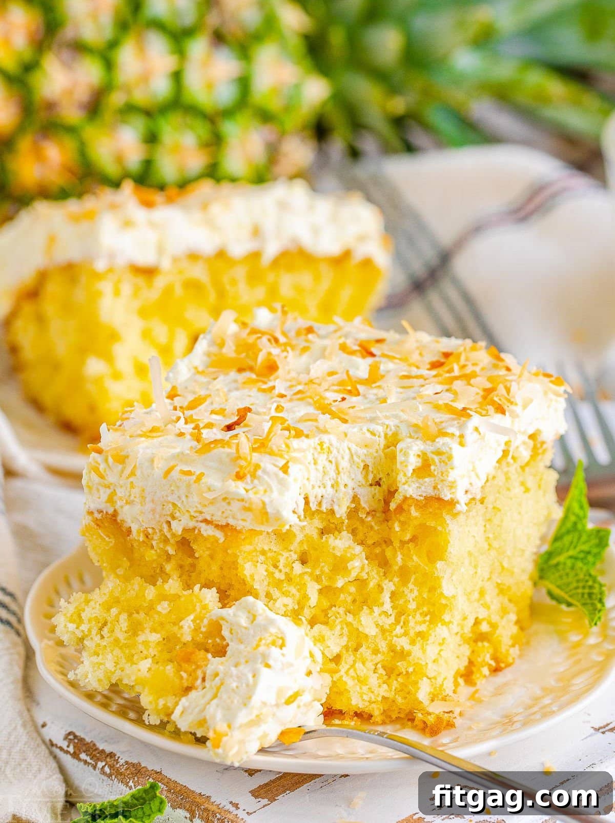 A delectable slice of Pina Colada Poke Cake sits on a white plate, with a fork gently removing a piece, highlighting the cake's moist interior and fluffy frosting. In the background, another slice and a whole, fresh pineapple enhance the tropical theme.