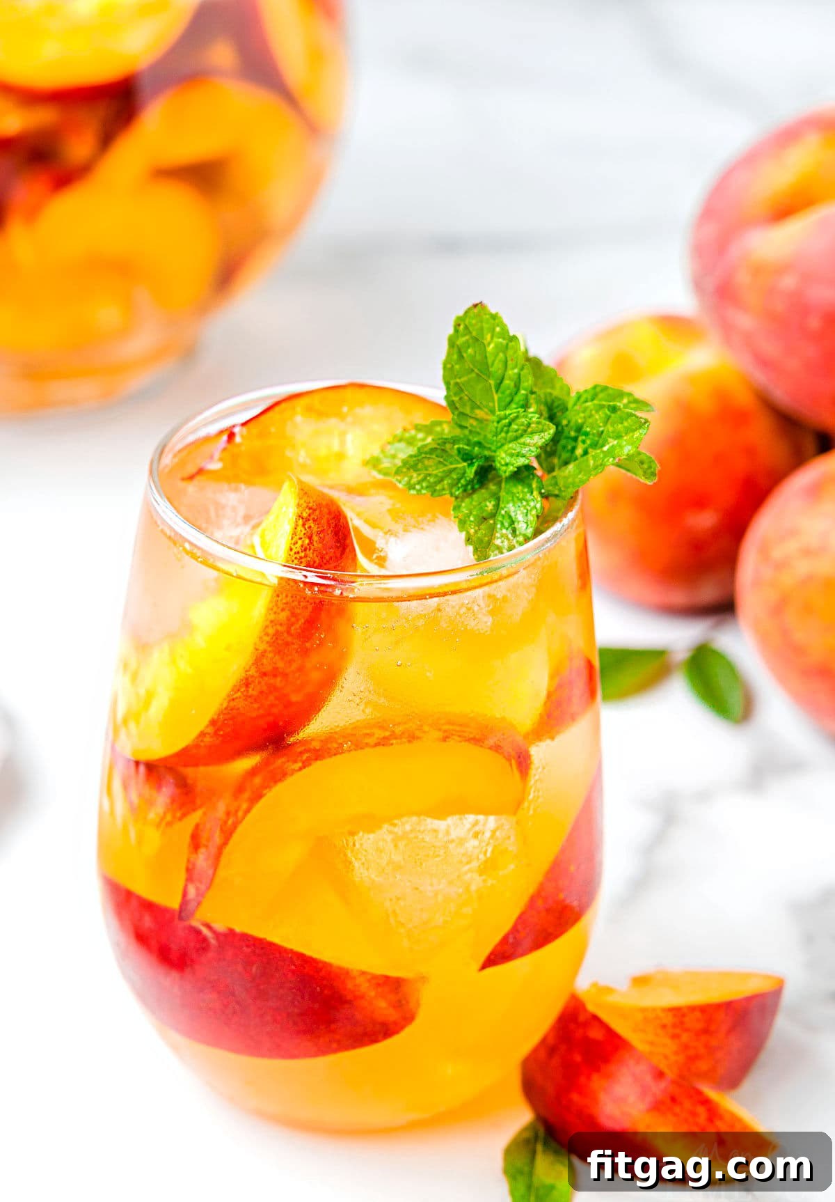Golden Peach Sangria 6 stemless wine glass filled with white sangria fresh peach slices and garnished with mint. fresh peaches to the back and the pitcher can be seen in the background.