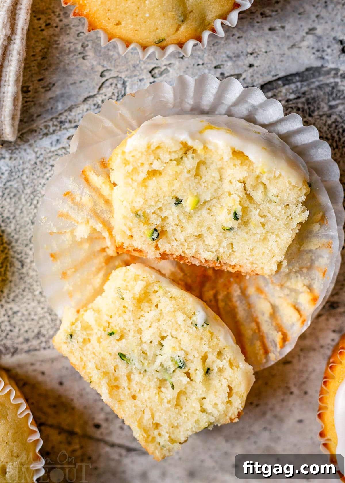 A perfectly baked lemon zucchini muffin in a white liner, sliced in half to reveal its tender, moist crumb, topped with a glistening lemon glaze and resting on a elegant gray backdrop. It highlights the texture and fresh ingredients.