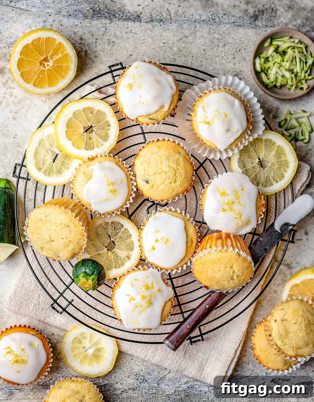 An overhead view of six perfectly glazed lemon zucchini muffins resting on a black wire cooling rack. Slices of fresh lemon are scattered around, emphasizing the citrus notes and freshness of the muffins.