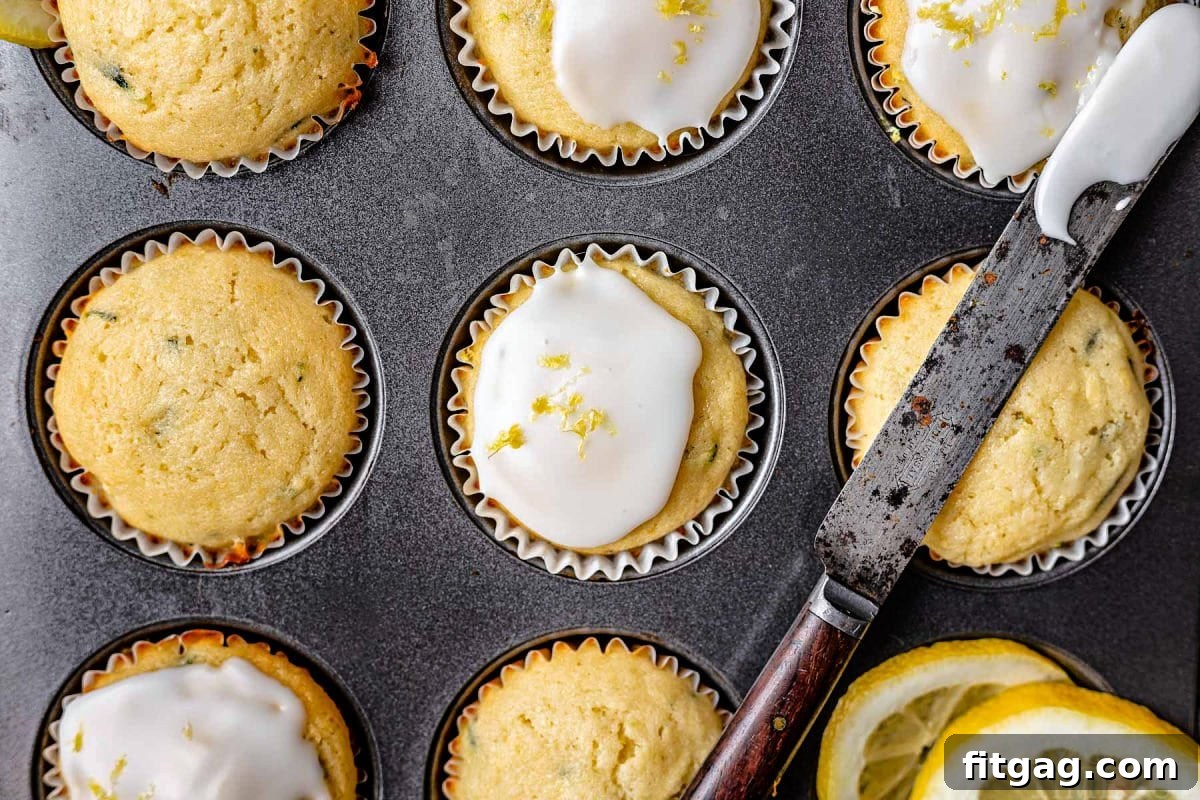 A muffin tin containing lemon zucchini muffins, with half of them beautifully glazed and a glazing knife resting across the top of the pan, ready for more.