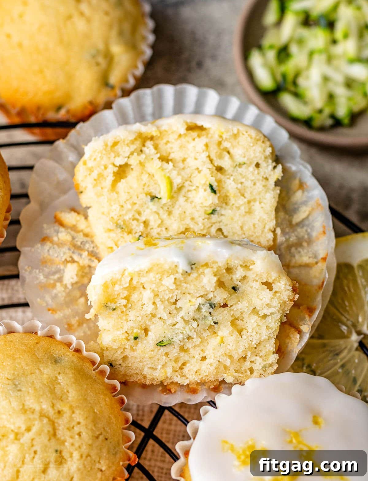 A close-up of a lemon zucchini muffin sliced in half, with one half resting on the other. Three whole muffins, some fresh lemon slices, and a small pile of shredded zucchini are artfully arranged nearby, showcasing the ingredients and final product.