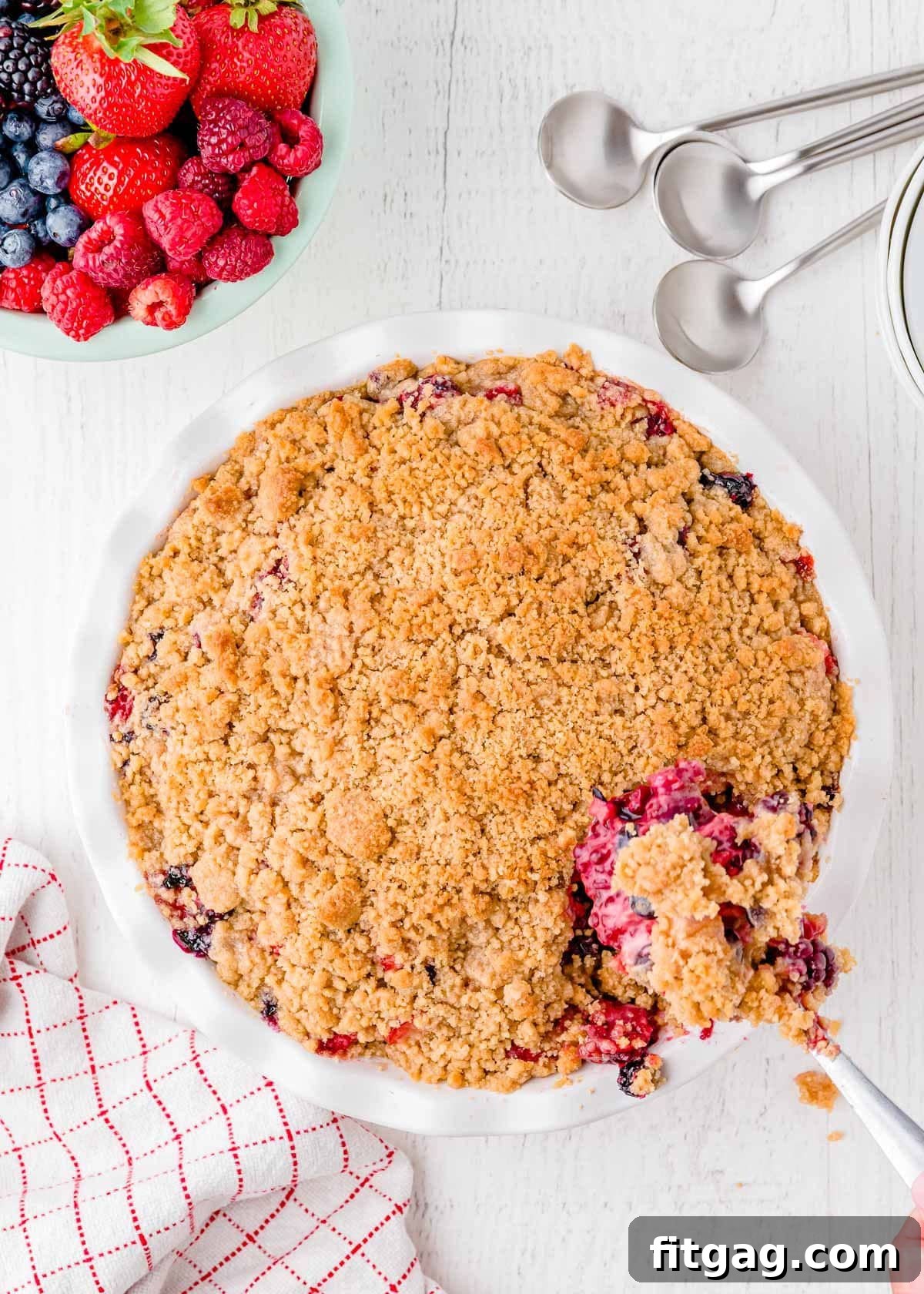 Top-down view of a berry crumble in a white pie dish, with a spoonful being removed, showcasing the bubbly fruit and golden topping.