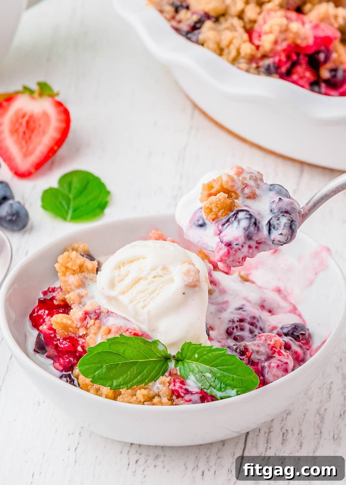 White bowl filled with berry cobbler topped with vanilla ice cream, with a spoonful of cobbler being scooped out.