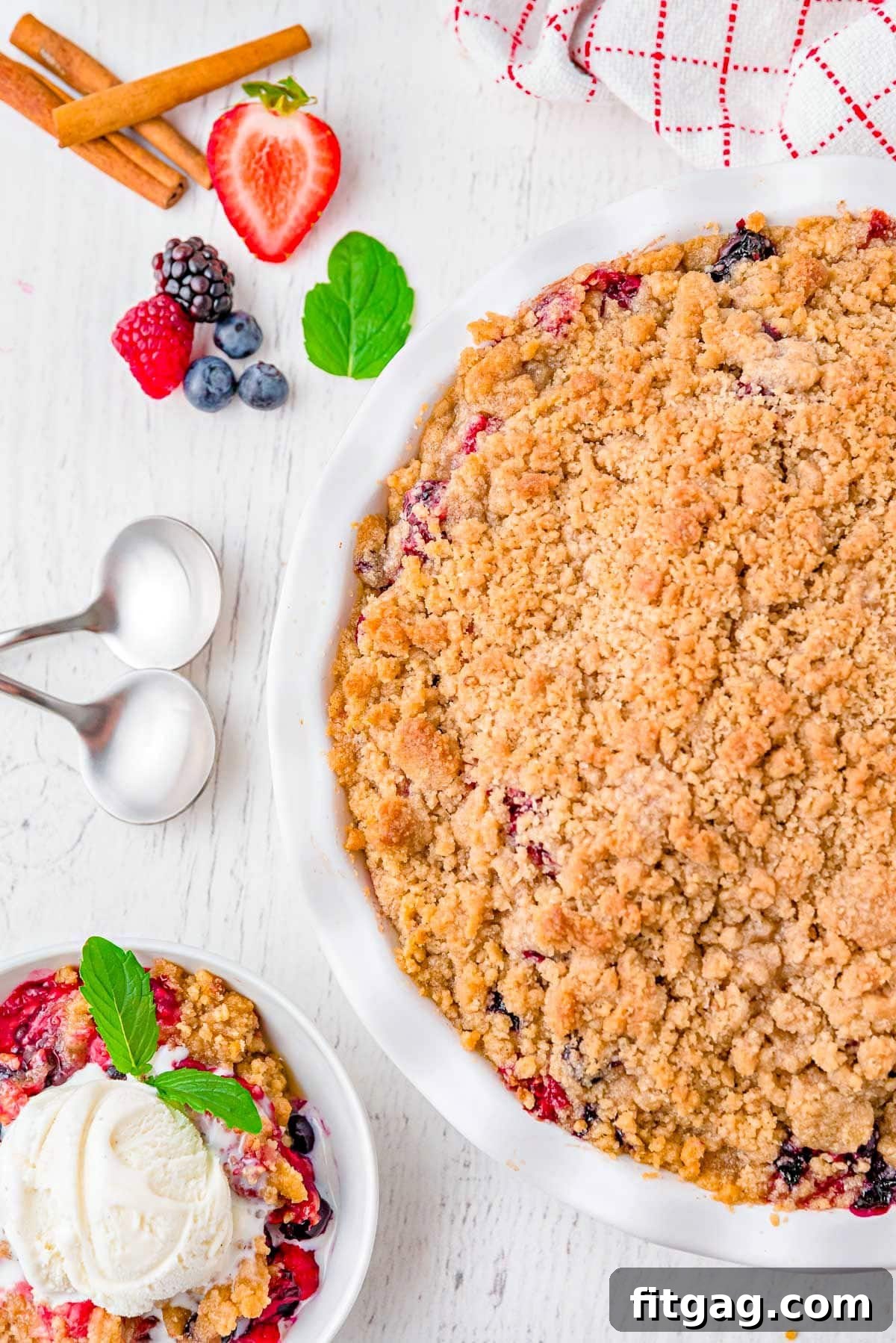 Top-down view of a berry crumble in a white pie plate with a serving removed and placed in a white bowl next to it. The crumble is generously topped with vanilla ice cream.