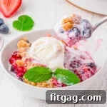 White bowl with berry cobbler in it topped with vanilla ice cream. spoonful of cobbler being scooped out.