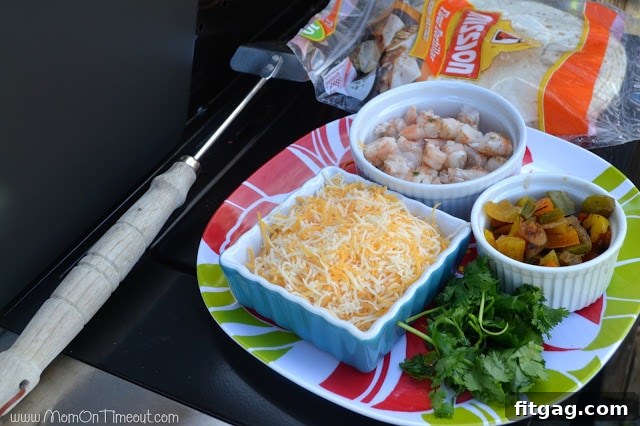 Variety of quesadilla ingredients like cheese, shrimp, cilantro, and tortillas laid out on a table next to a grill