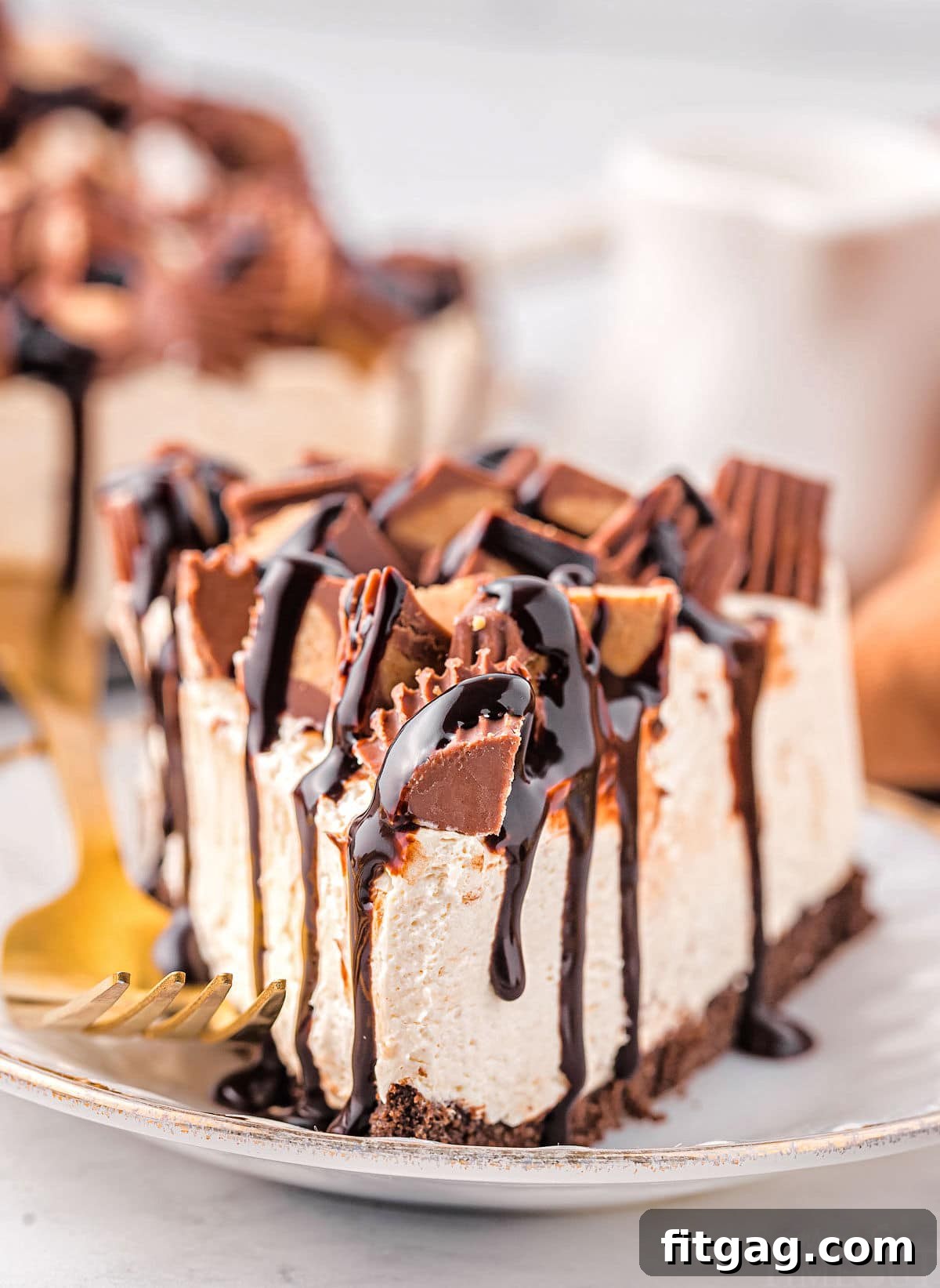 A delectable slice of no-bake peanut butter cheesecake, generously topped with Reese's candies and hot fudge, resting on a small gold-rimmed white plate with a gold fork beside it. The perfect blend of creamy, sweet, and nutty flavors.