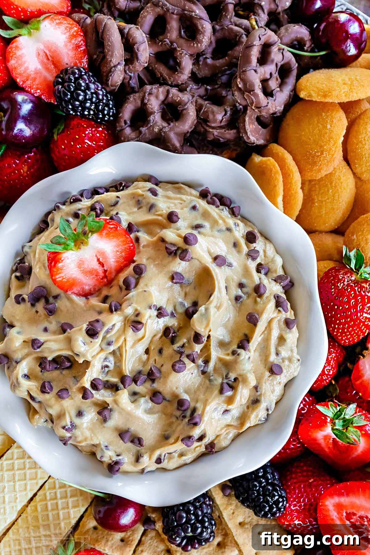 Decadent Cookie Dough Dip 2 A top-down view of a white bowl overflowing with creamy chocolate chip cookie dough dip, surrounded by an inviting assortment of fresh strawberries, chocolate-dipped pretzels, Nilla wafers, and other vibrant fruits and cookies, perfect for dipping.
