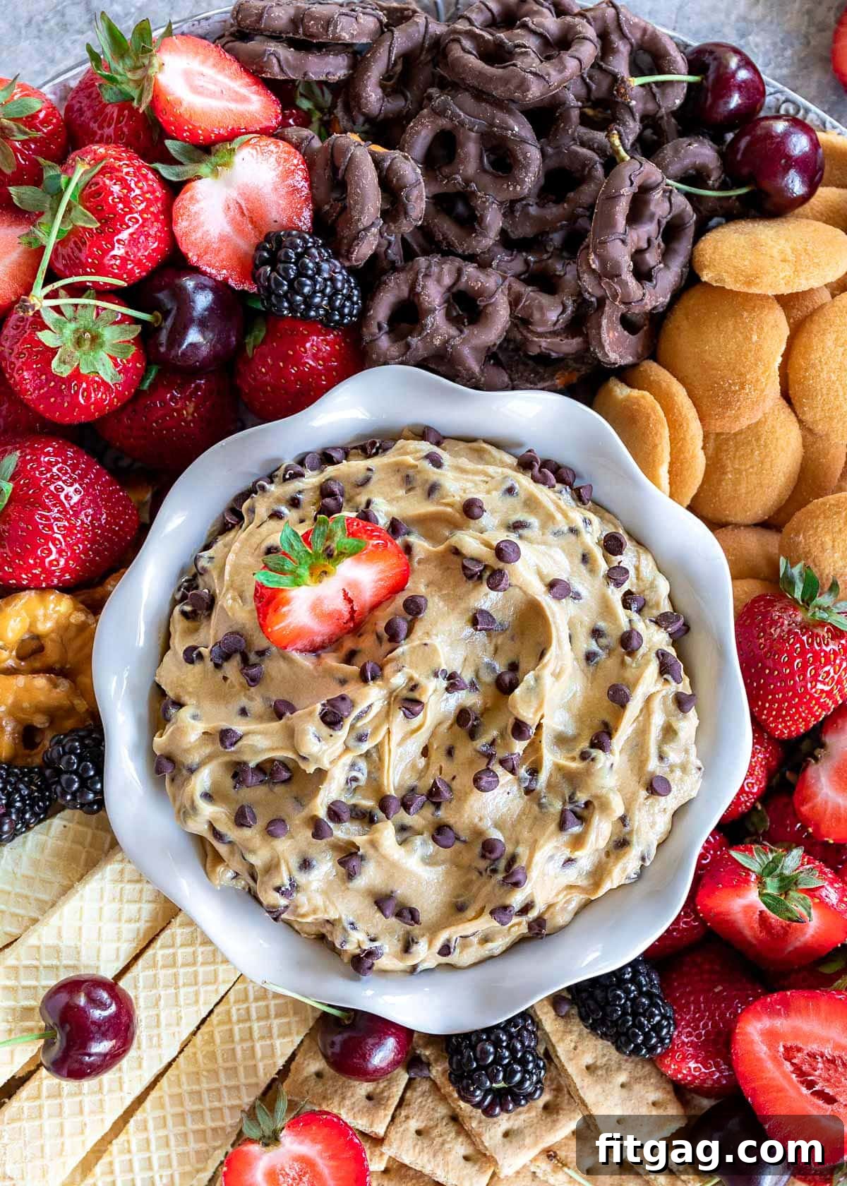 Decadent Cookie Dough Dip 4 A mesmerizing top-down perspective of creamy chocolate chip cookie dough dip nestled in a white bowl, artfully surrounded by an abundant assortment of fresh strawberries, decadent chocolate-dipped pretzels, classic Nilla wafers, and other vibrant fruits and crispy cookies.