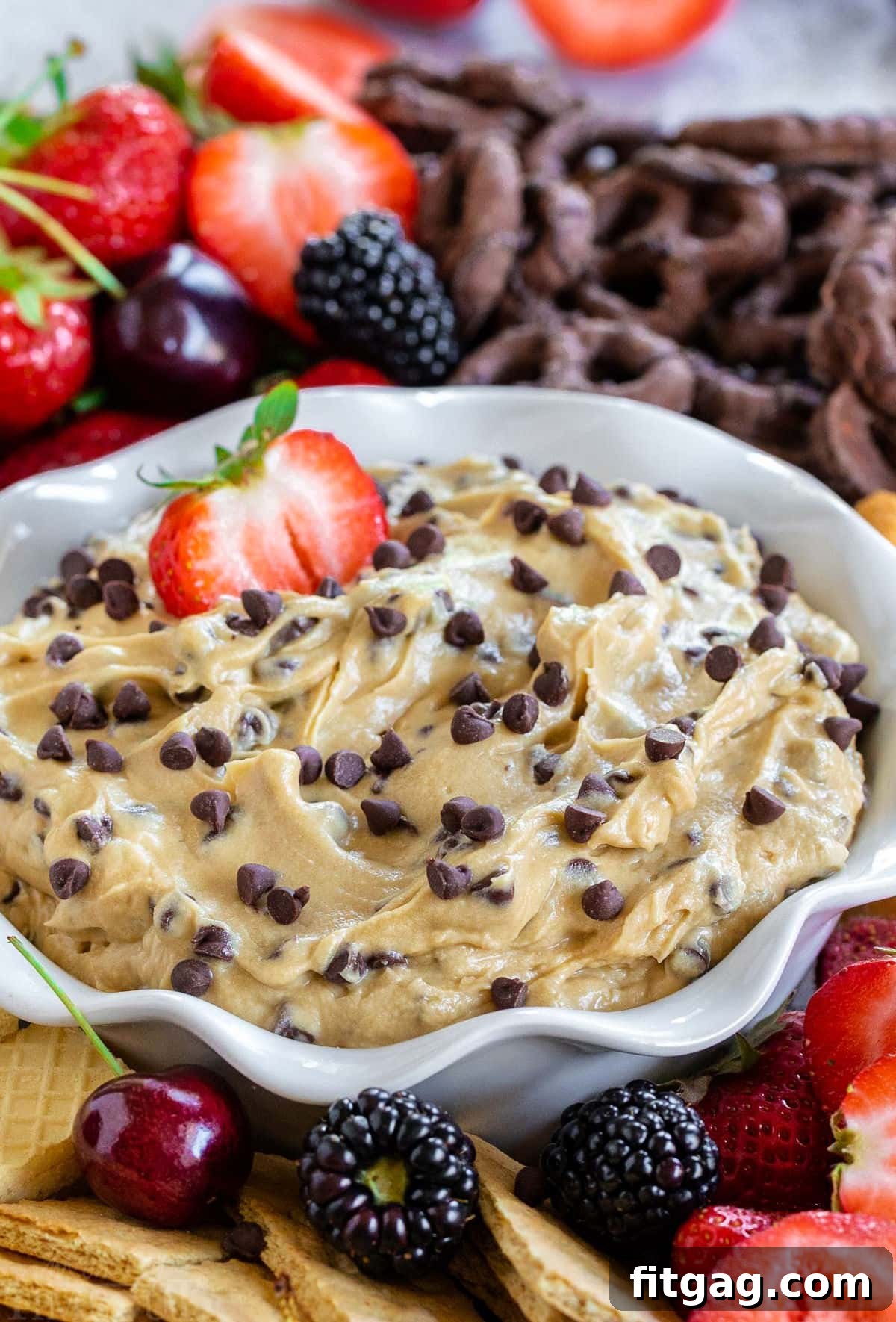 Decadent Cookie Dough Dip 5 A charming bowl with fluted edges holds a generous portion of creamy chocolate chip cookie dough dip, with a half-eaten strawberry playfully dipped into its rich texture. The bowl is artfully surrounded by an assortment of vibrant fresh fruit and crisp cookies, creating an inviting spread.