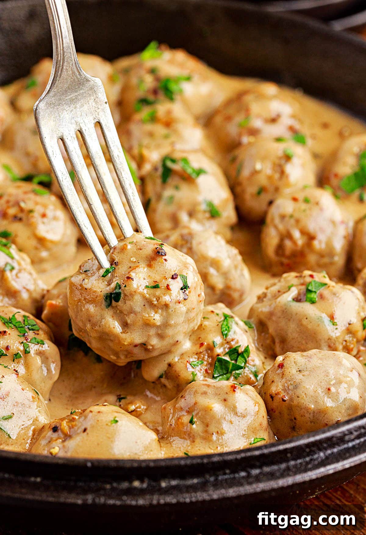 A fork picking up a Swedish meatball from a cast iron skillet filled with creamy sauce and meatballs, highlighting the delicious texture and rich gravy.