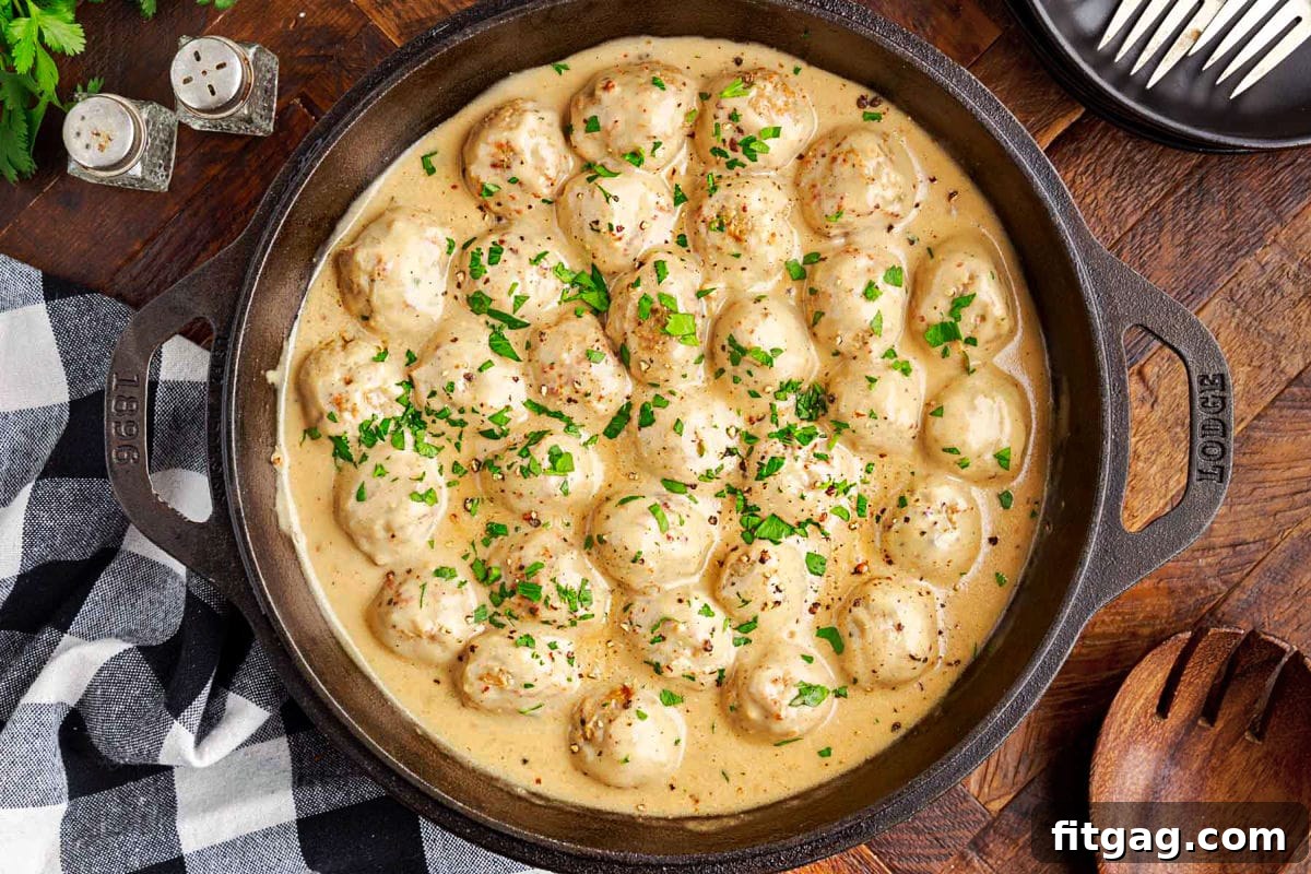 A wide shot looking down into a black cast iron skillet brimming with Swedish meatballs and creamy gravy, resting on a dark wood surface with a black and white checked napkin nearby, evoking a rustic, cozy feel.
