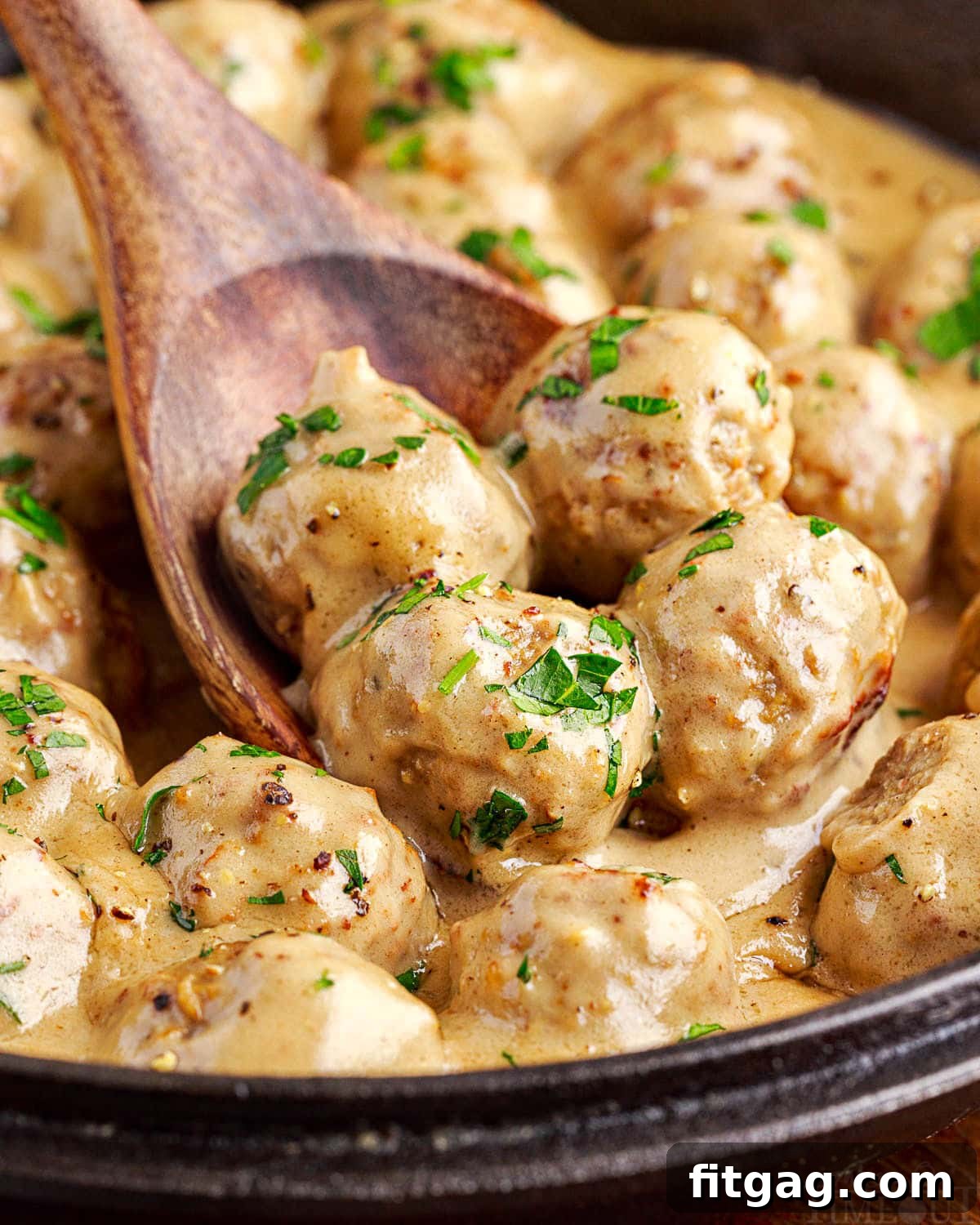 Four perfectly browned Swedish meatballs on a wooden spoon, being scooped out of a skillet filled with the remaining meatballs and creamy sauce, ready to be served.