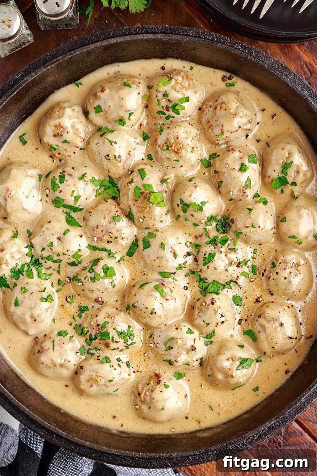 A top-down view of a black cast iron skillet filled with creamy Swedish meatballs and gravy, resting on a dark wood surface with a black and white checked napkin nearby, ready to be served.