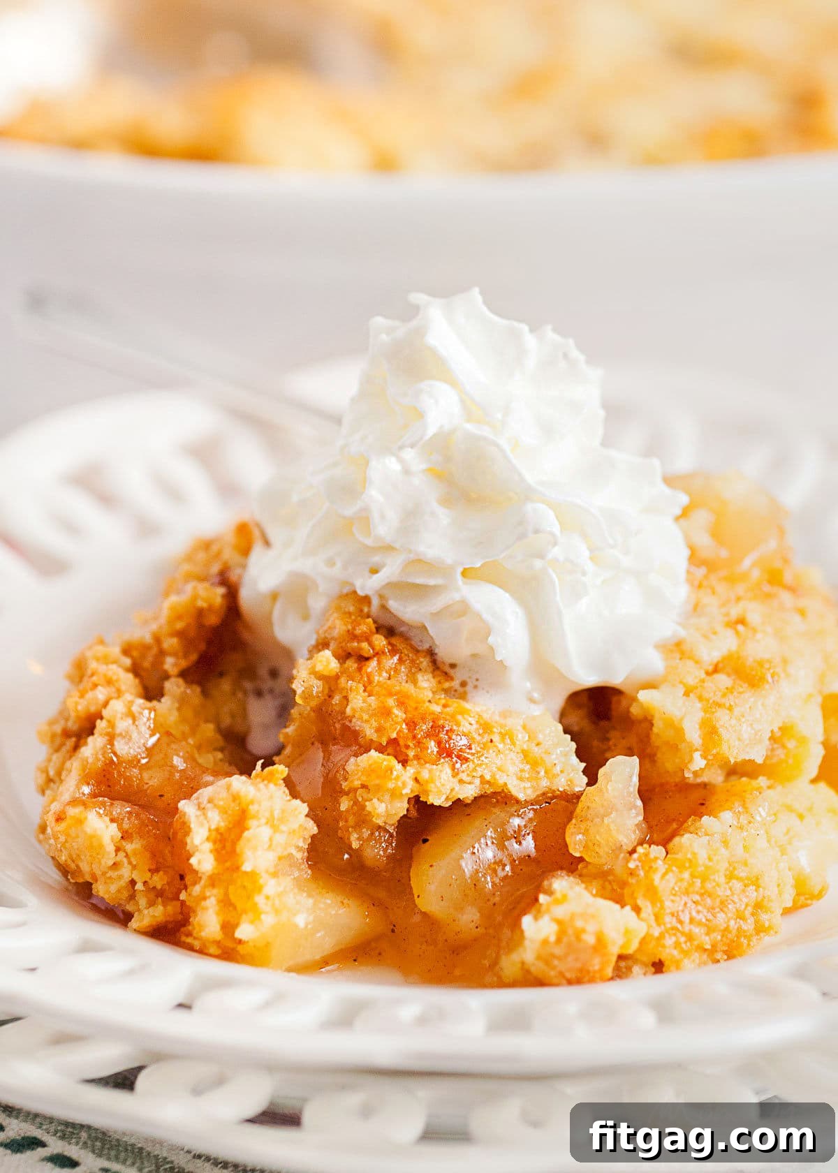 No-Fuss Apple Cobbler 2 A generous scoop of golden-brown easy apple cobbler, topped with a swirl of fresh whipped cream, served on a white lace-edged plate. In the blurred background, the white casserole dish holds the remaining warm cobbler, ready for more servings.