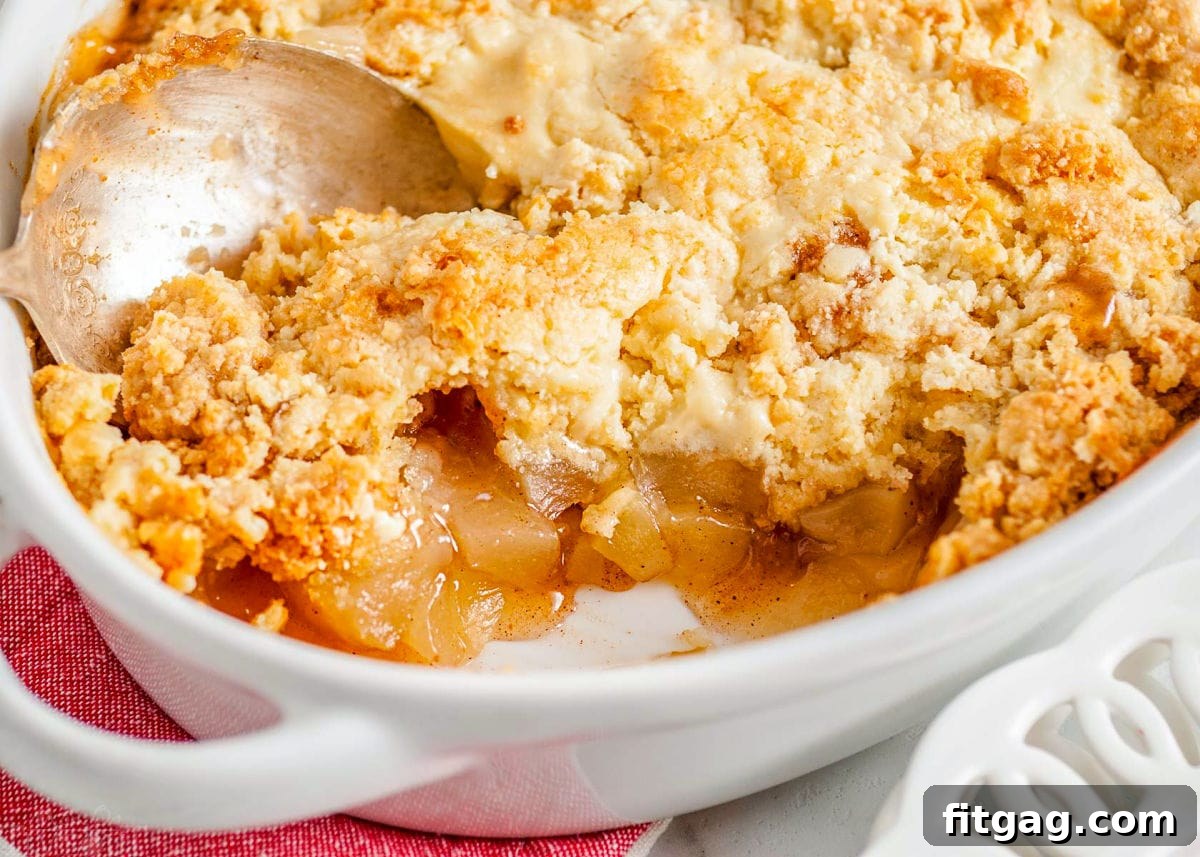 No-Fuss Apple Cobbler 4 A top-down view of a freshly baked easy apple cobbler in a white baking dish. Several portions have been removed, showcasing the tender apple filling and the golden cake mix crust. An antique-style serving spoon rests invitingly in the dish.