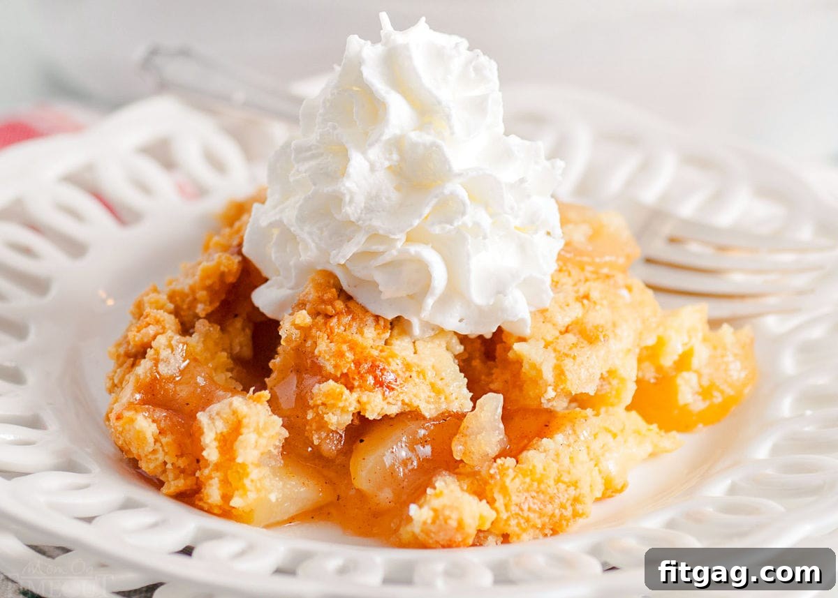 scoop of apple cobbler served on a white lace edged plate topped with fresh whipped cream. white casserole dish with remaining cobbler in the background.