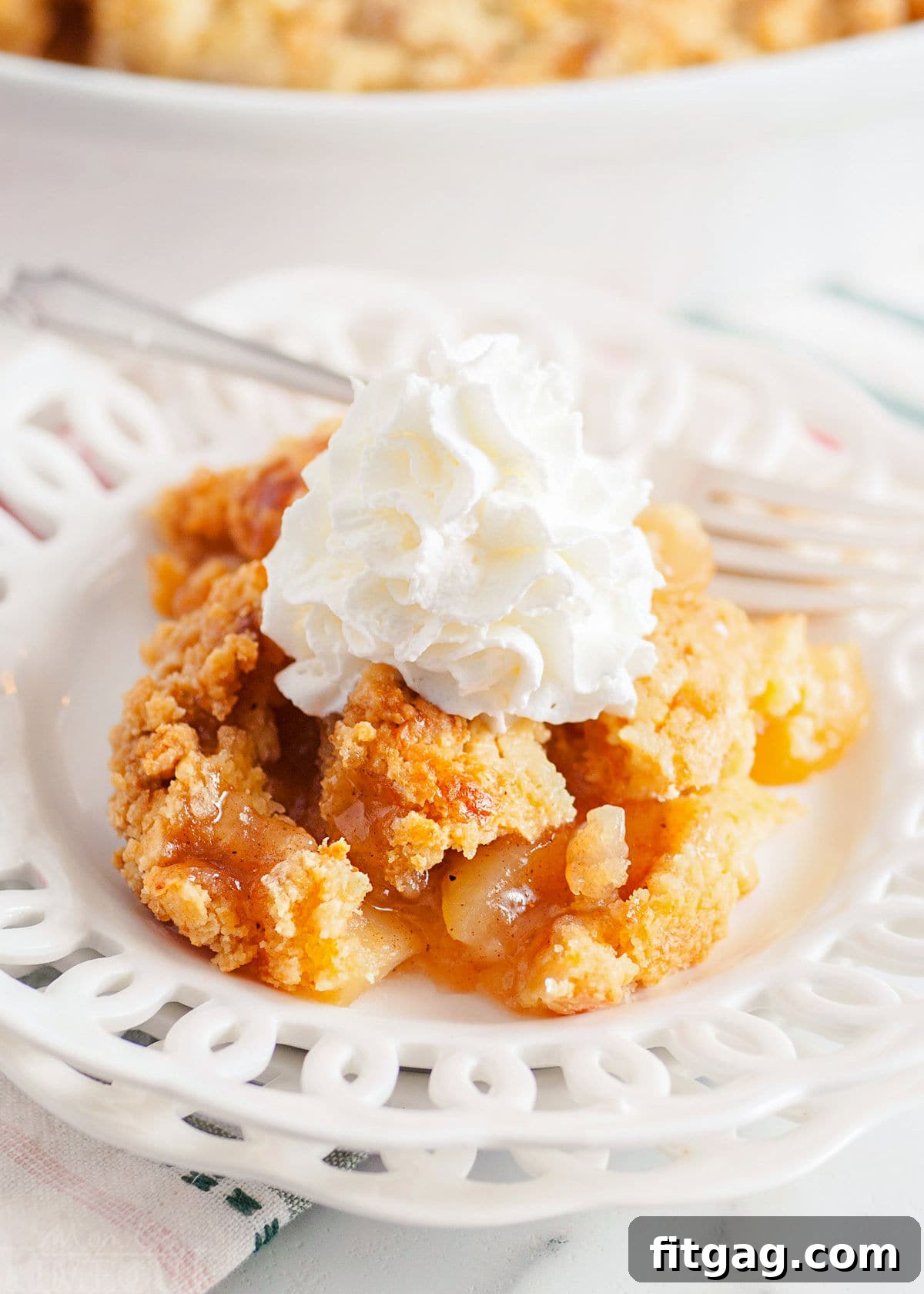 No-Fuss Apple Cobbler 8 A close-up of a serving of easy apple cobbler on a white lace-edged plate, generously topped with whipped cream, with the baking dish holding the remaining cobbler in the soft background.