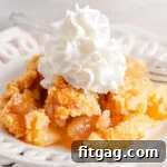 No-Fuss Apple Cobbler 9 A scoop of golden-brown apple cobbler, crowned with creamy whipped cream, on a white plate. The baking dish with more cobbler is visible in the background.