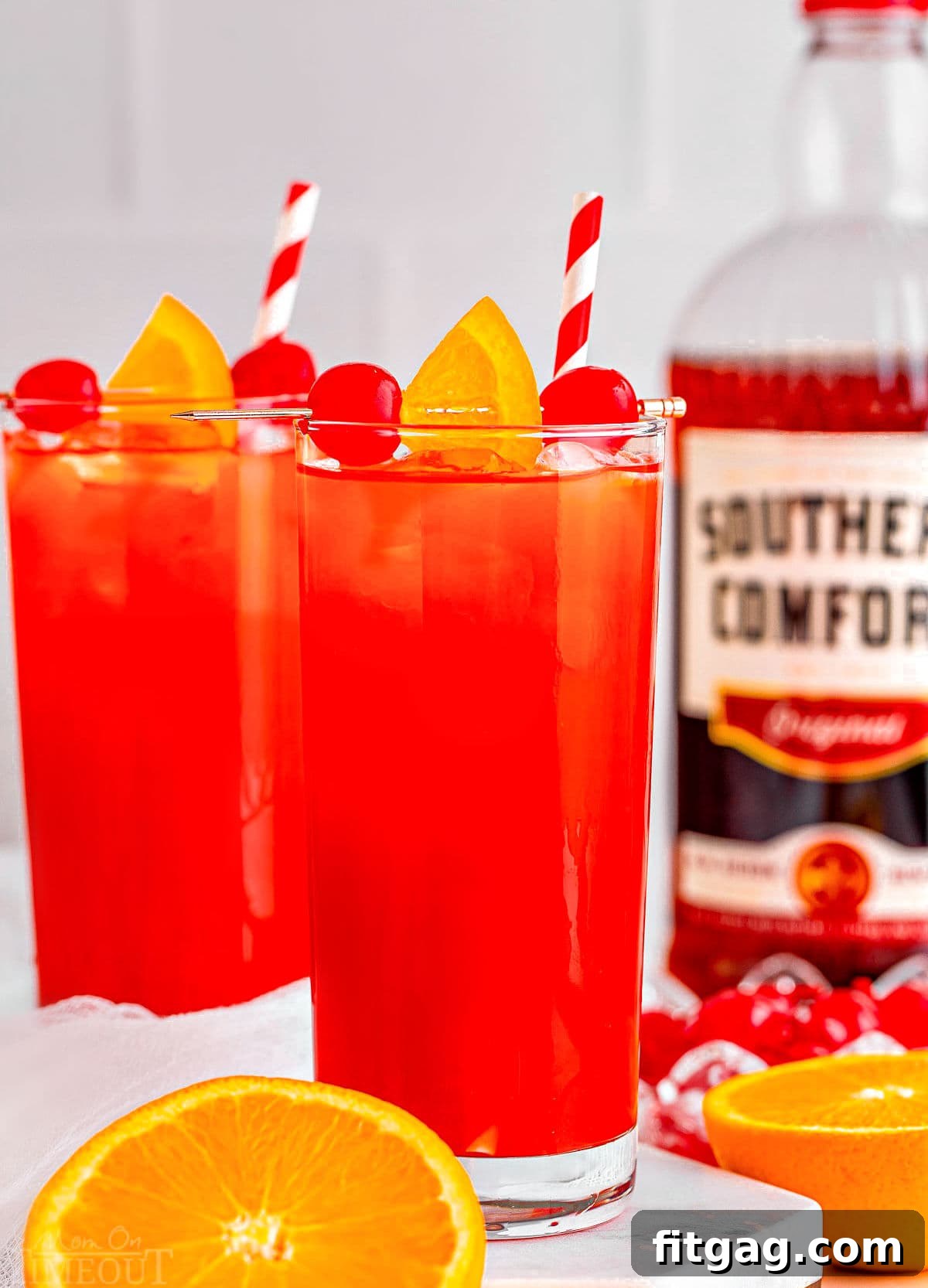 two tall glasses filled with alabama slammer recipe with red and white striped straws. bottle of southern comfort in the background and one orange has been cut in half.