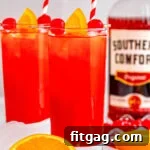 two tall glasses filled with alabama slammer recipe with red and white striped straws. bottle of southern comfort in the background and one orange has been cut in half.