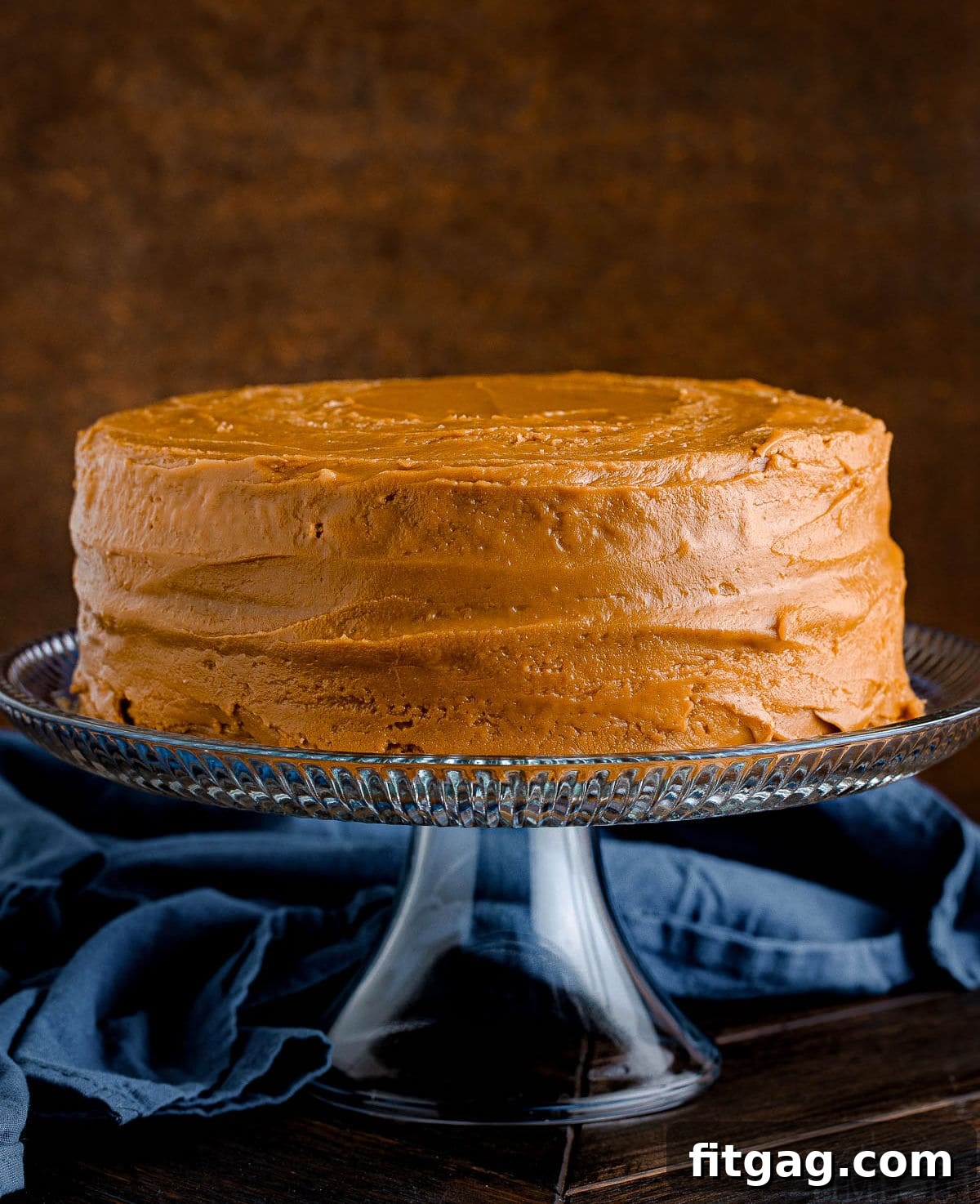 Two-layer caramel cake with rich caramel frosting, presented elegantly on a clear glass cake stand with a soft navy blue napkin in the background. A perfect centerpiece for any gathering.