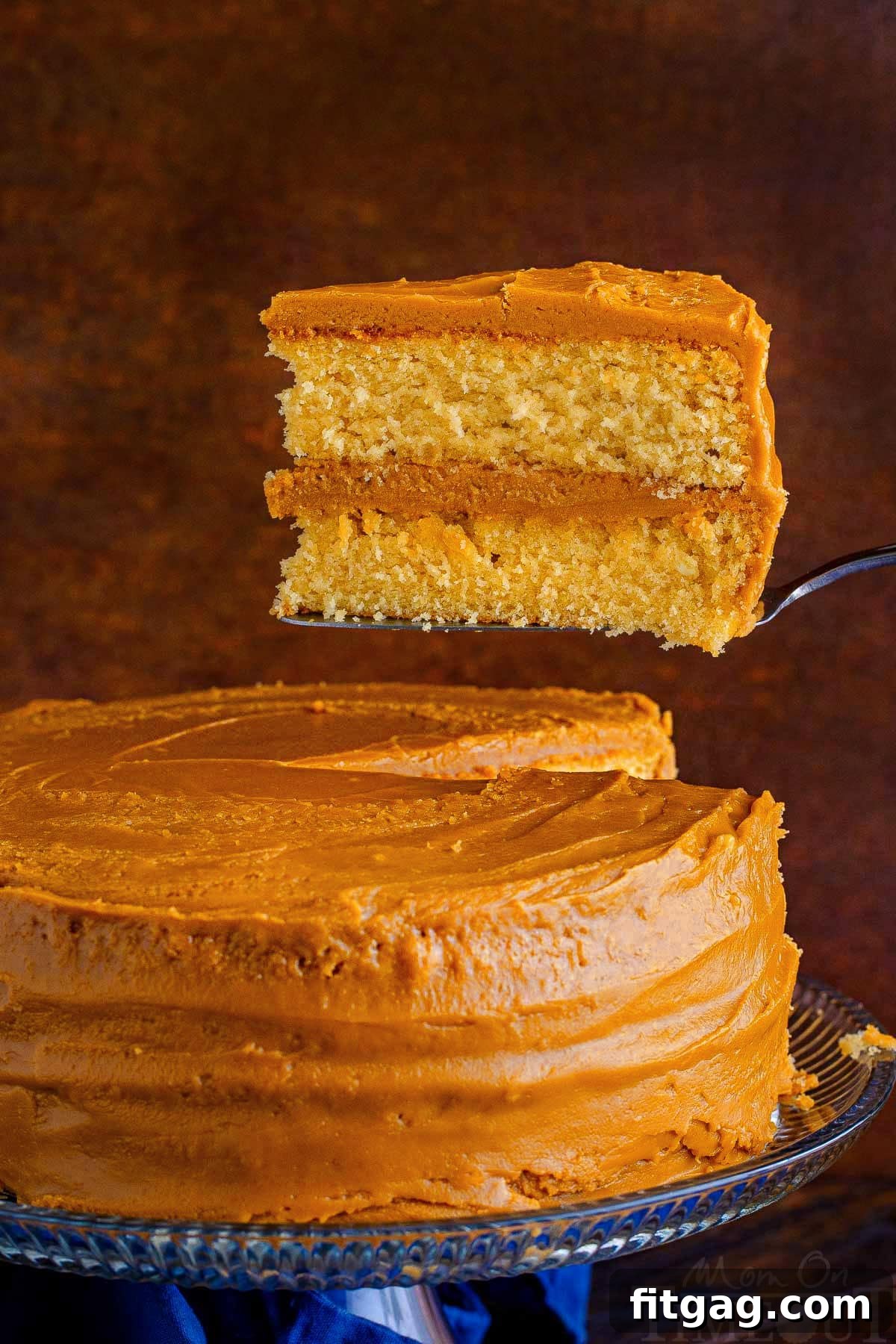 A large slice of decadent caramel cake, held aloft and slightly pulled away from the main cake on a stand, showcasing its moist layers and thick frosting.