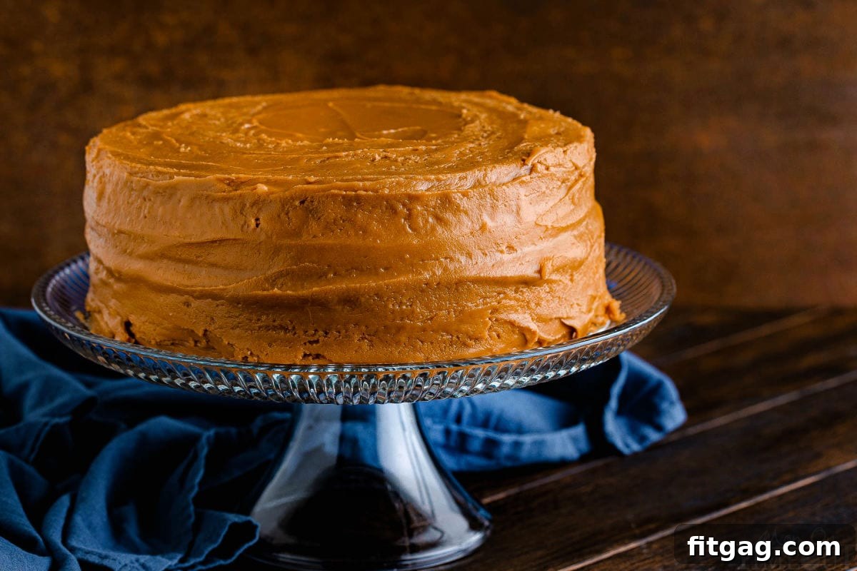 A beautifully frosted two-layer caramel cake sitting elegantly on a clear glass cake stand, with a rich navy blue napkin creating a sophisticated backdrop.
