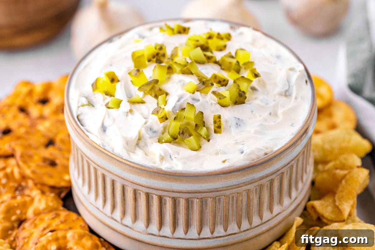 Creamy Dill Pickle Dip 4 small decorative gray clay bowl with ridged detailing is filled with pickle dip and topped with more diced pickles. pretzels and chips surround the bowl.