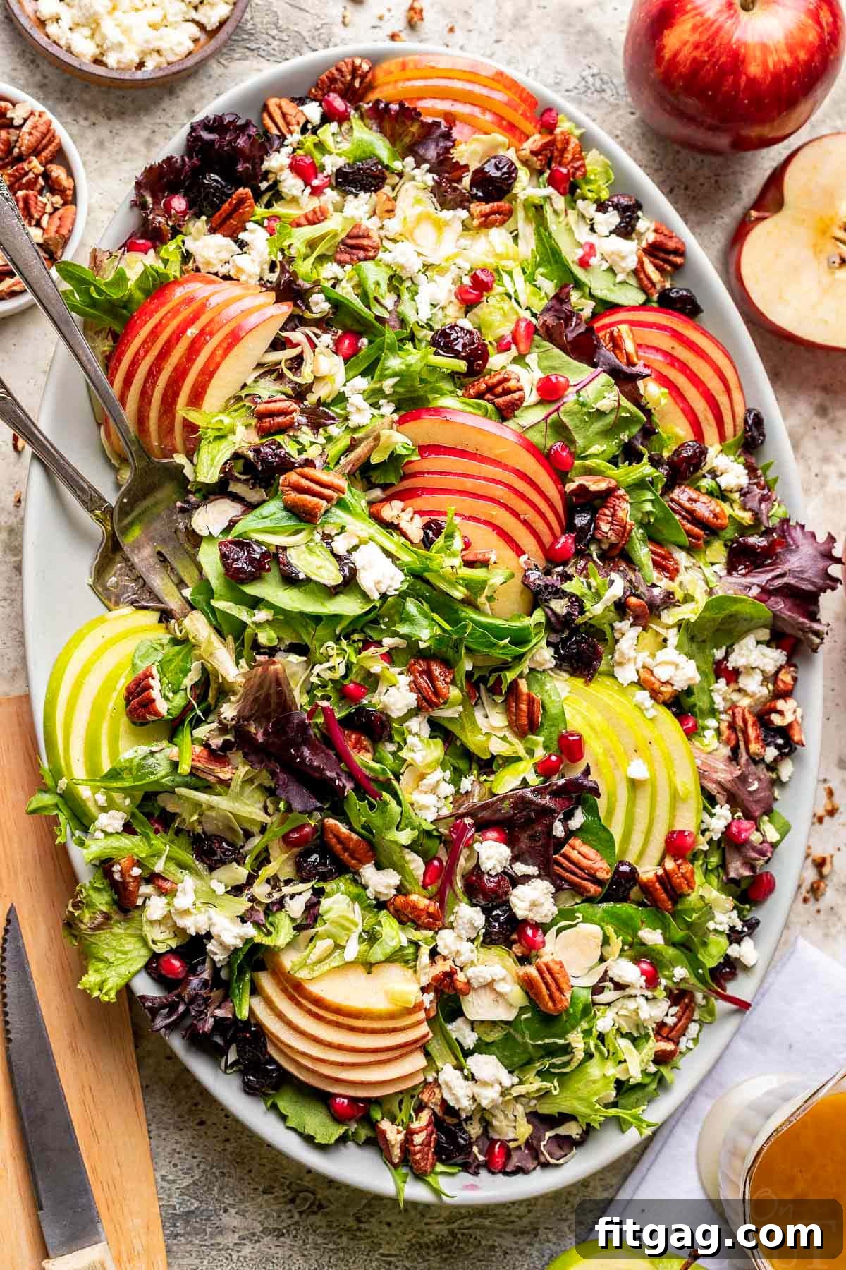 top down look at apple salad made with brussels sprout, apples, cranberries and topped with a vinaigrette. oval serving bowl is sitting on a gray surface with cut apples around it.