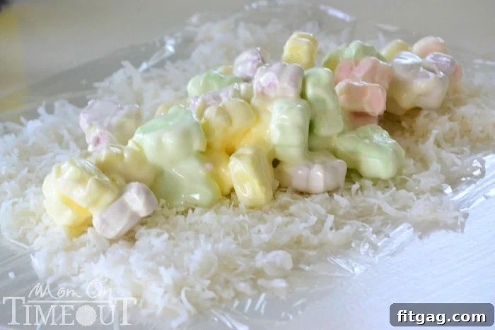 Melted CandiQuik and colorful marshmallows spread down the center of a bed of coconut on plastic wrap, ready to be rolled.