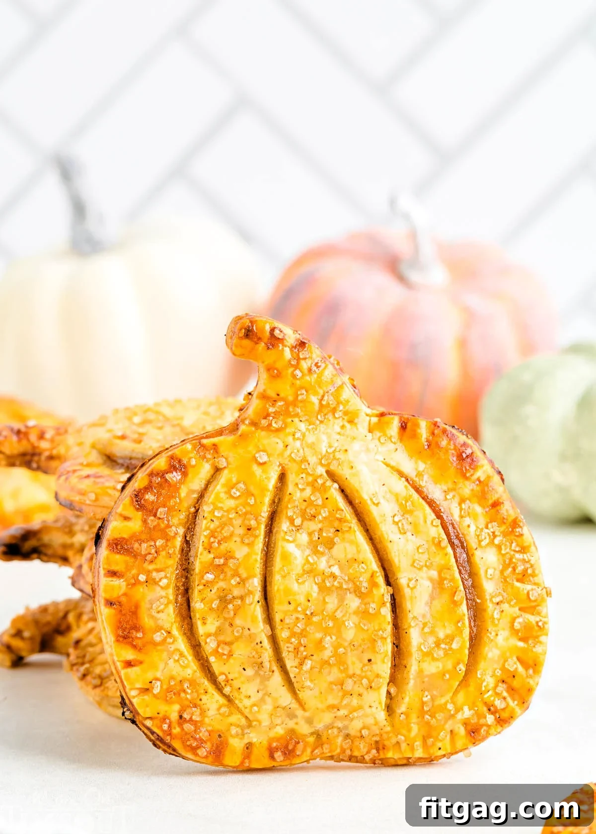 Pumpkin Pie Pockets 2 A single pumpkin-shaped pumpkin hand pie standing upright in front of a stack of similar pies. In the background, an assortment of decorative pumpkins hints at the autumn season.