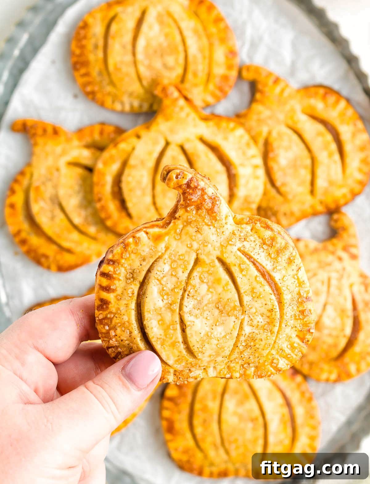 Pumpkin Pie Pockets 5 A hand holds up a single, perfectly baked pumpkin-shaped hand pie over a tray filled with many more, showcasing their inviting golden-brown crust.