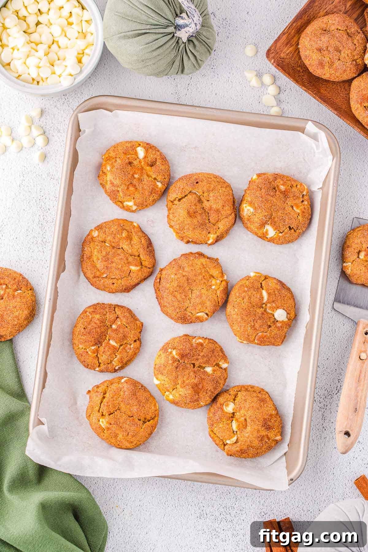 A top-down shot of freshly baked pumpkin snickerdoodle cheesecake cookies arranged on a baking sheet, with a green napkin peeking into the corner, and additional cookies scattered around, ready to be enjoyed. The scene evokes a cozy, autumnal baking aesthetic.