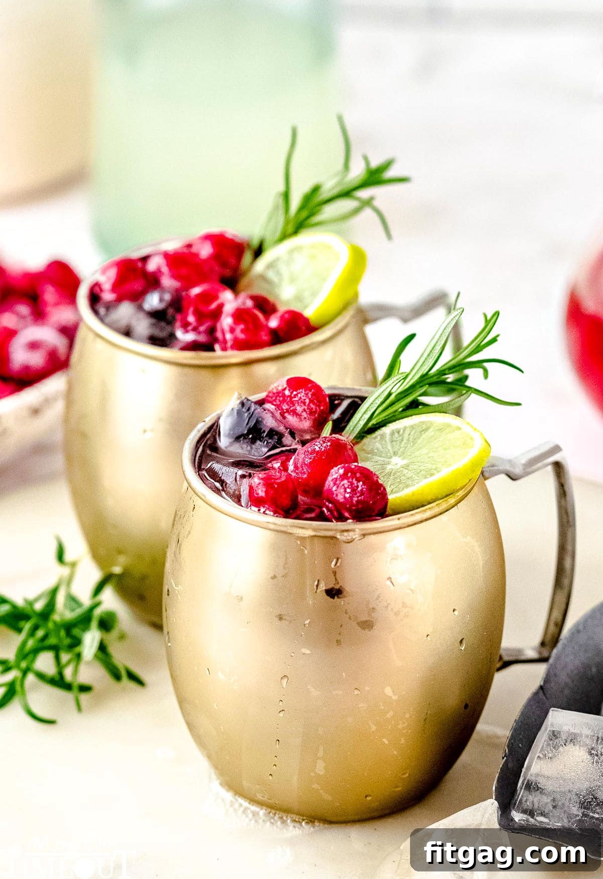 Crimson Moscow Mule 2 Two vibrant Cranberry Moscow Mule cocktails adorned with fresh cranberries, lime slices, and a sprig of rosemary, set on a clean white surface.