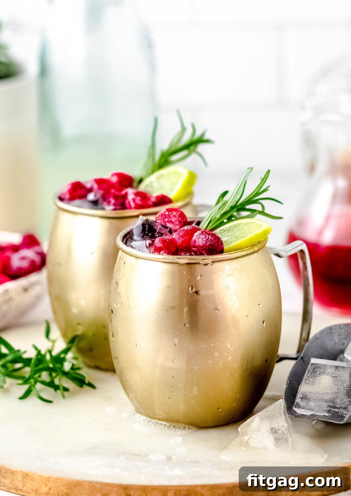 Crimson Moscow Mule 6 Two Cranberry Moscow Mule cocktails garnished with cranberries, lime slices, and a sprig of rosemary, presented on a rustic white board.
