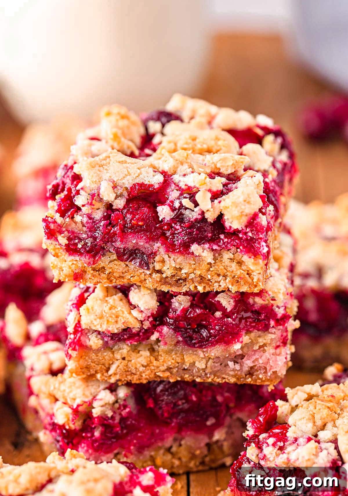 Three cranberry crumb bars stacked on each other on a dark wood board with a glass of milk in the background, highlighting the layers and texture.