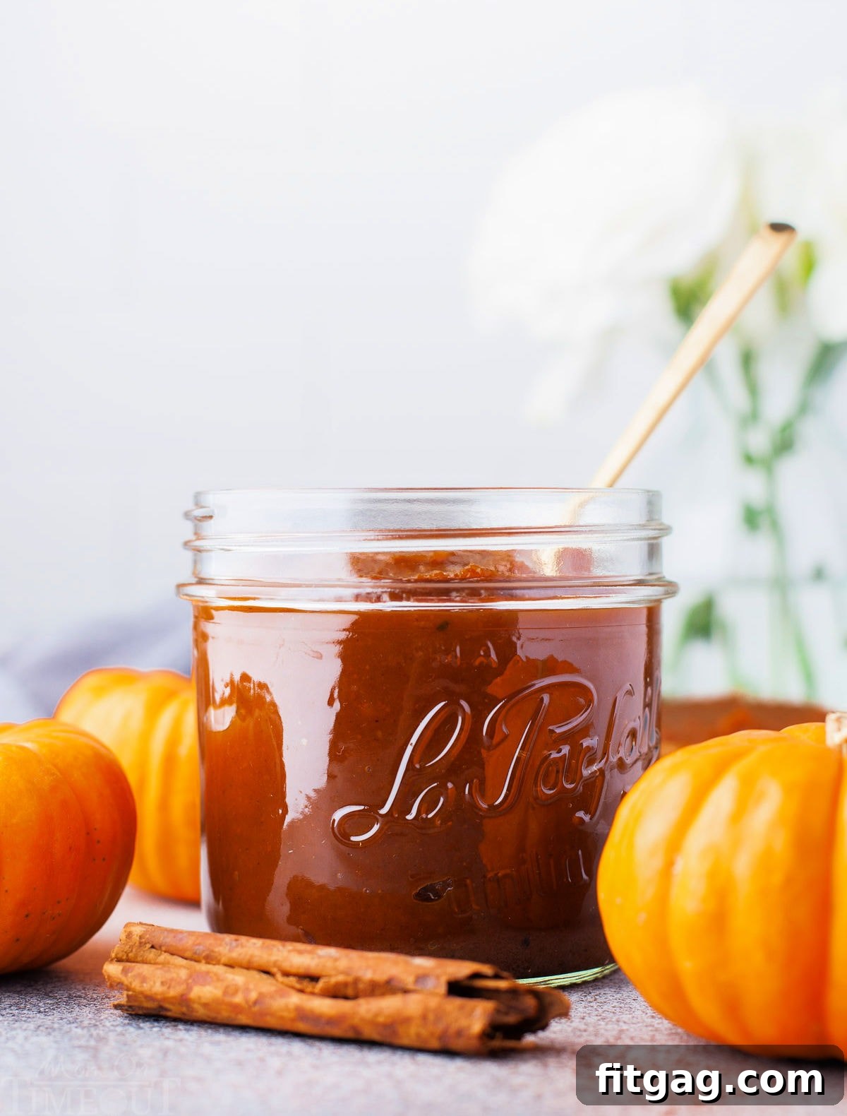 front view of glass jar filled with homemade pumpkin butter. three small decorative pumpkins and a cinnamon stick are sitting around the jar.