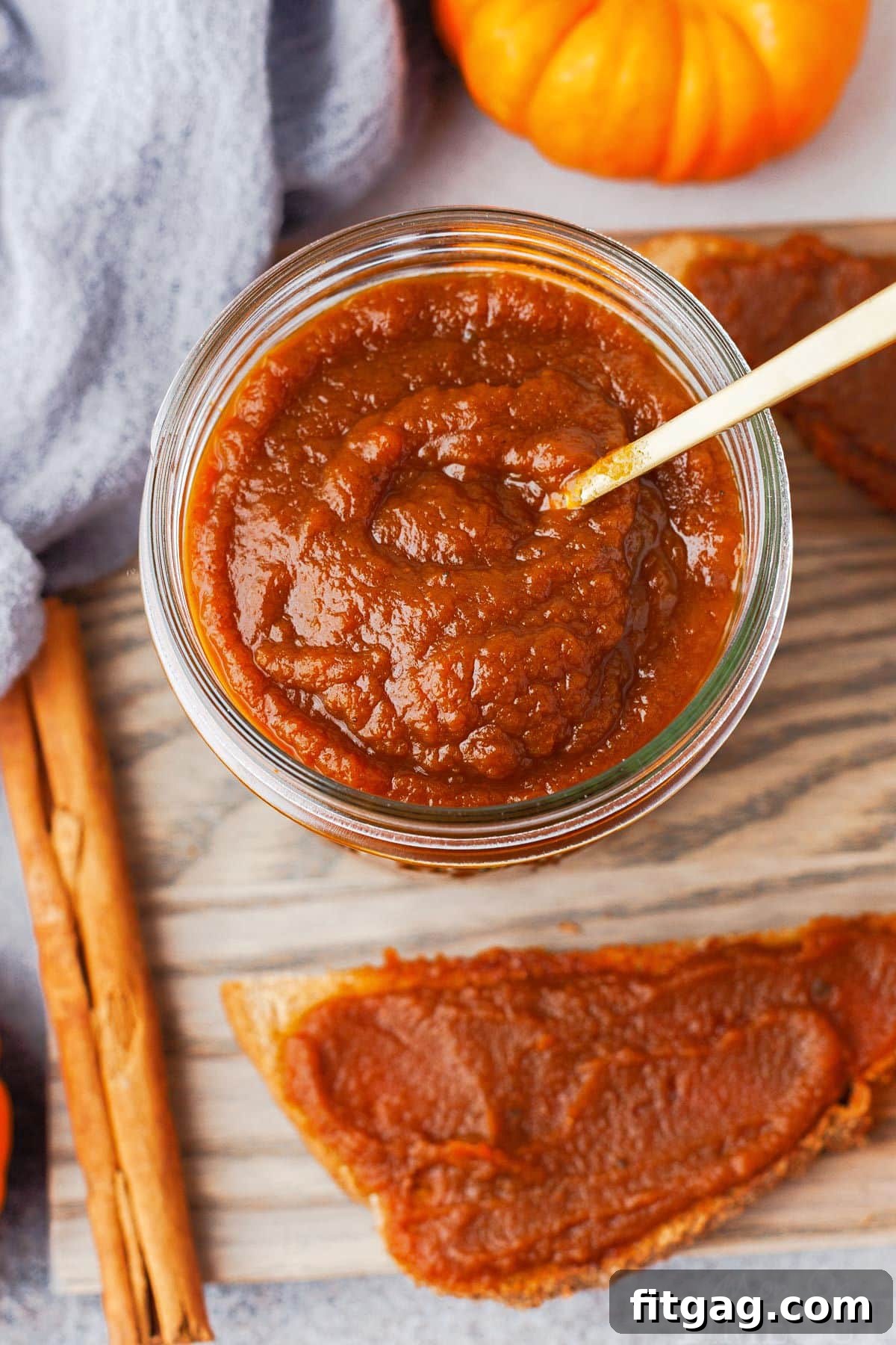 top down view of jar of pumpkin butter with small gold spoon sitting on a gray board with a half piece of toast with pumpkin butter slathered on it.