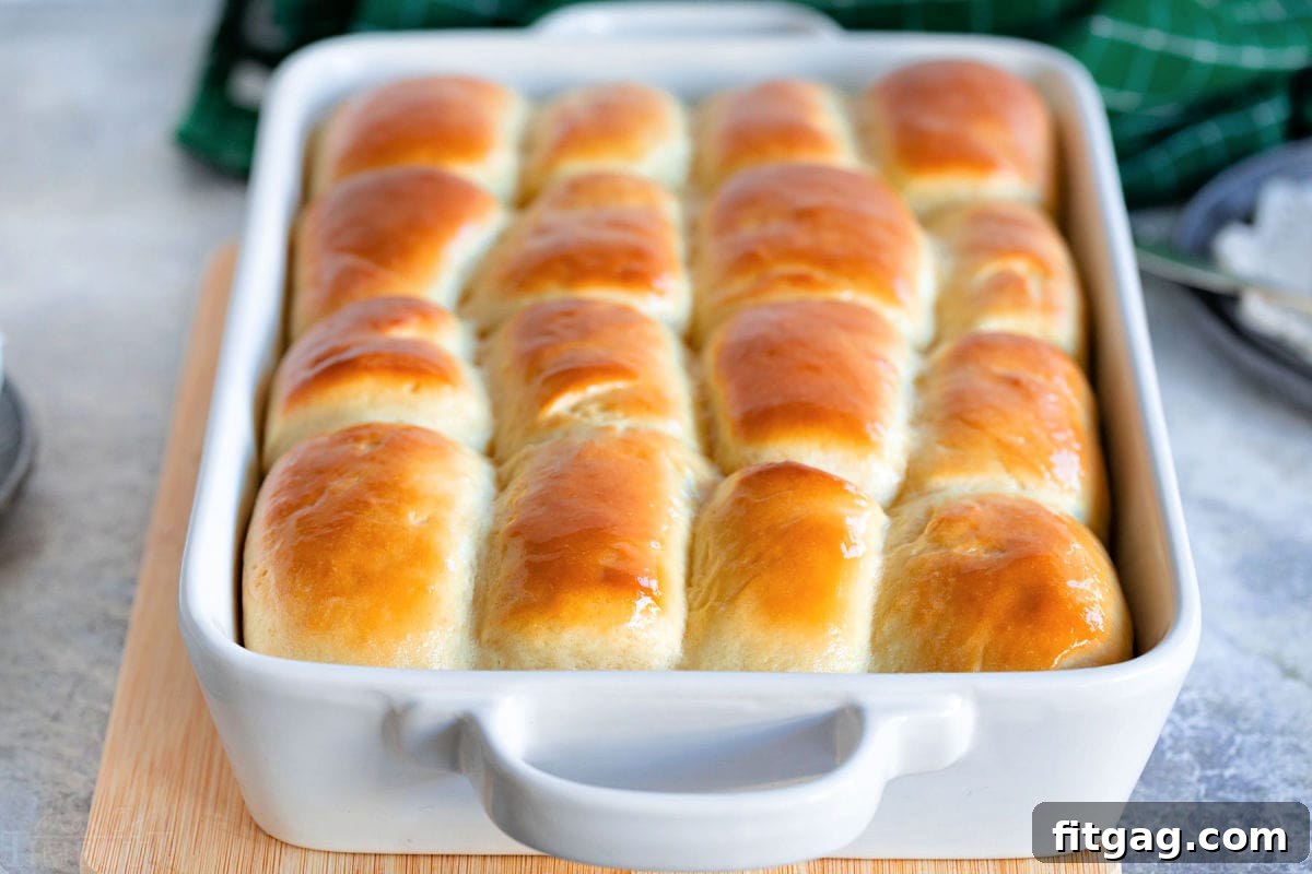 white baking dish with golden brown parker house rolls ready to be enjoyed. baking dish is sitting on cutting board.