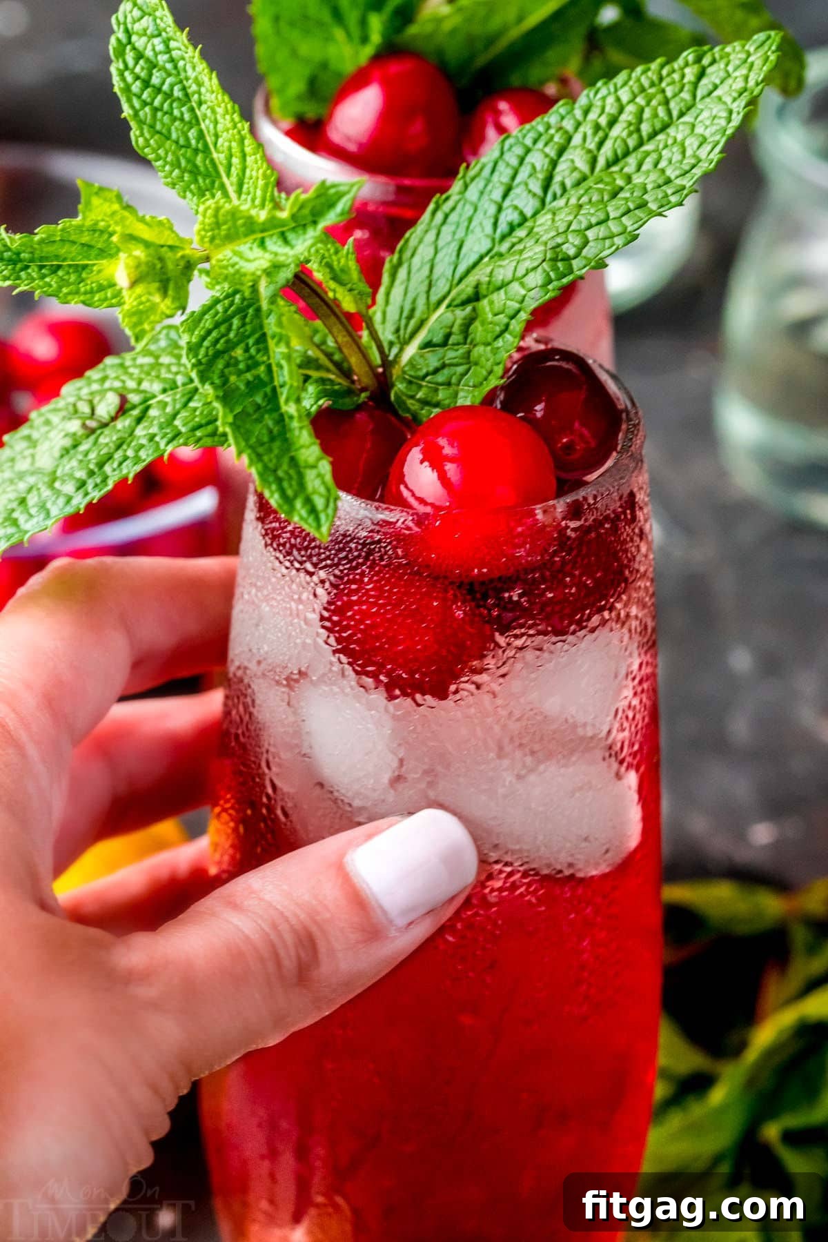 Crimson Gin Sparkler 4 A hand gracefully holds a Cranberry Gin Cocktail, topped with a fresh mint sprig, against a softly blurred festive background.